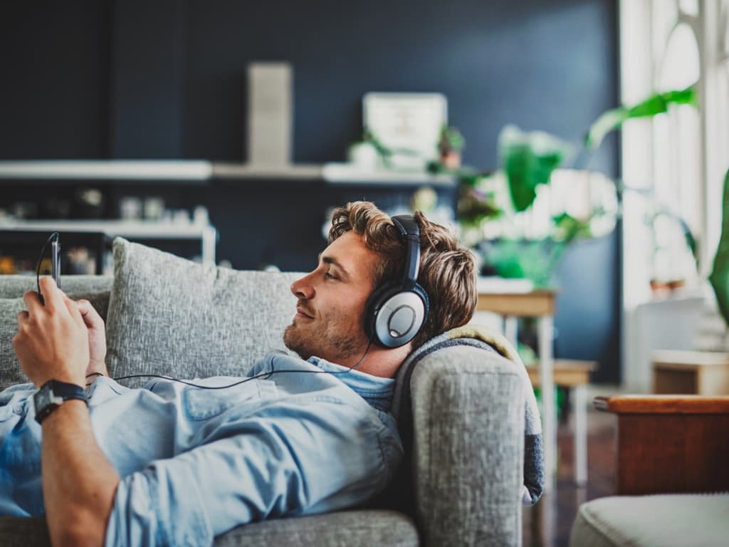En mand sidder på en sofa med høretelefoner på, fokuseret på sin telefon, mens han slapper af
